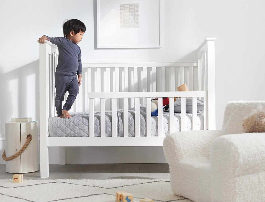 Shop Pottery Barn Kids — Kids' and Nursery Furniture, Baby Gifts and Kids' Decor | Featuring toddler boy in blue pajamas standing in Kendall Crib in Simply White with Toddler Bed Conversion, with grey quilted bedding.